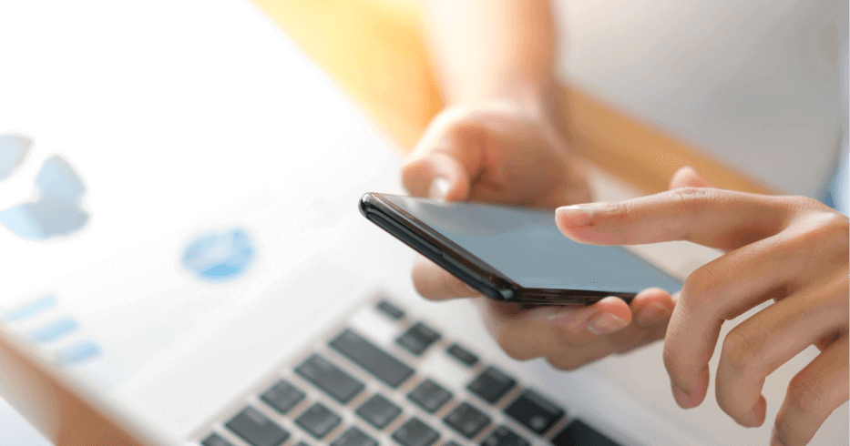 Image of a person using a cell phone while simultaneously typing on a laptop keyboard